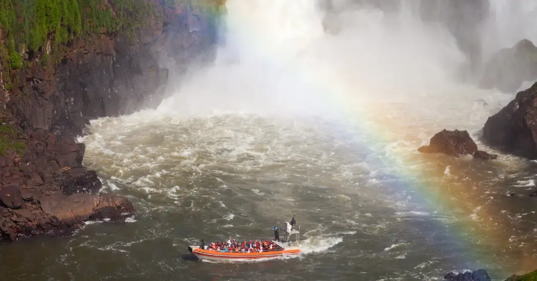 Tourists on the boat ride exploring the Argentinian side of the Iguazu Falls with a beautiful rainbow appearing in the sky.